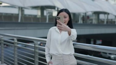 Güzel Gülümseyen Kız Sokakta Duruyor ve Arkaplanda Köprüsü Olan Selfie Çekiyor. Charming Positive Woman in Business Sutire Outside Hafıza için bir video kaydetti. Halk ve Teknoloji