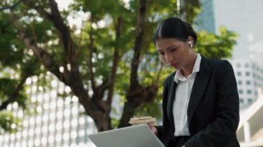 Laptopuyla dışarıda çalışırken iş yerinde atıştırmakla meşgul Asyalı bir kadın. CEO, bilgisayarındaki uzak görevler sırasında sandviç yiyor. Halk ve Teknoloji Konsepti
