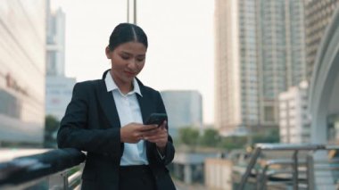 Dışarıda duran ve telefonuyla çalışan, ofis kıyafetleri giymiş bir kadın. Olumlu Asyalı Yönetici, müşteri soruşturmalarına akıllı telefonuyla cevap veriyor. Halk ve Teknoloji Konsepti