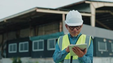 Depoya yakın bir yerde duran erkek mühendis üniforma giyerken iş için tablet kullanıyor. Kıdemli işçi çalışıyor ve internetten not alıyor. İnsan ve İş Konsepti