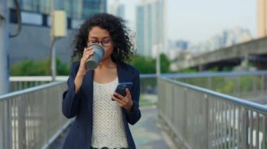 Güzel iş kadını işten sonra yürüyor, telefonunu tutarken kahve içiyor ve iş sohbetini okuyor. Charming Ofis çalışanı kahve molasında. Akıllı telefonunu kullanıyor ve şehirde geziniyor.