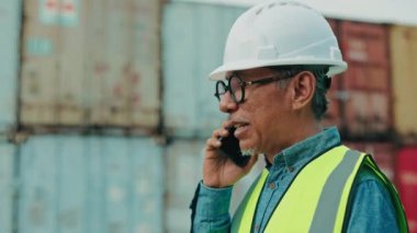 Sert şapka ve yansıtıcı yelek giyen üst düzey bir Asyalı işçi açık gökyüzünün altındaki bir nakliye konteynerinde telefon görüşmesi yapıyor..