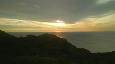Tayland 'da bulutlu gün batımında sakin okyanus suyuna karşı uçurum silueti. Altın güneş ışığı Phuket 'te huzurlu bir tatil için rahatlatıcı atmosfer yaratıyor.