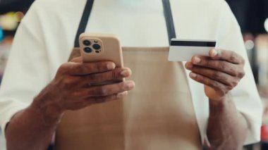 Elinde akıllı telefon ve kredi kartı olan önlüklü Afro-Amerikan bir adamın elleri. Erkek garson ödeme bilgilerini cep telefonundan giriyor. Guy servis çalışanı dijital aktarımı tamamlar
