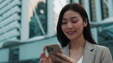 Ofis binasının yanında duran akıllı telefon kullanan mutlu Taylandlı kadının portresi. Saçı gevşek, cep telefonuyla internette gezinen Asyalı olumlu bir iş kadını. Şehir Caddesinde kadın müdür