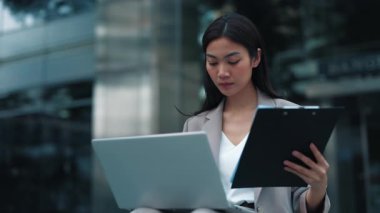 Taylandlı kadın portresi iş belgelerinden bilgisayara veri girişi yapıyor. Şehir caddesinde oturuyor. Konsantre Asyalı esmer iş kadını elinde pano olan şirket belgelerini gözden geçiriyor.