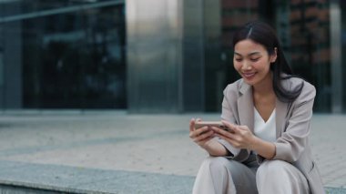 Taylandlı iş kadını, ara verildiğinde ofis binasının yanındaki merdivenlerde oturup telefonda komik videolar izlerken güler. Neşeli Asyalı kadın sosyal medyada olumlu içeriğe sahip.