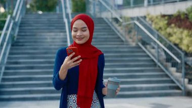 Şehir merkezinde yürüyen akıllı telefon ve kahve fincanı olan Malezyalı bir iş kadınının portresi. Genç Müslüman kadın tatilde iş sohbetini okur. İş profesyoneli