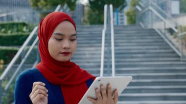 Taş basamakların yanındaki tablet bilgisayarda interneti kaydıran Malezyalı kadın şirket çalışanı portresi. Türbanlı kadın iş adamı şehir merkezinde duran e-postaları okuyor.