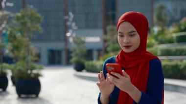 Akıllı telefonlu Malezyalı kadının portresi parkta online piyango kazanmasını kutluyor. Neşeli Müslüman kadın, cep telefonuna bakıp gülümseyerek ve hareket ederek mutluluğunu ifade ediyor.