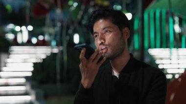Siyah tişörtlü mutlu Asyalı cep telefonu hoparlörüyle konuşuyor ve gece caddesinde durup gülerek gülüyor. Duygusal adam son olumlu haberi paylaşmak için ses gönderiyor.