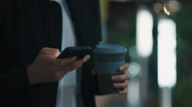 Erkek kişinin akıllı telefon kullanırken eko bardakla içecekle el kaldırmasını yakın çekim. Gece vakti.