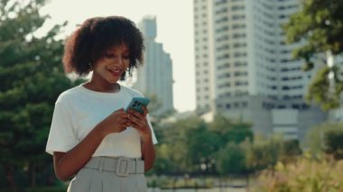 Parkta gökdelenlere karşı akıllı telefondan mesaj gönderen mutlu kadın portresi. Afro-Amerikalı kadın, şehir ortamında cep telefonuna bakarken gülümsüyor. Şehir yaşam tarzı