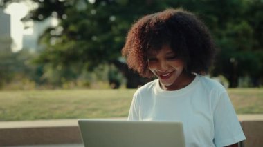Parkta dizüstü bilgisayarda yazı yazan mutlu Afrikalı Amerikalı kadın portresi. Sapık saçlı bir bayan yönetici bilgisayarla ağaçların karşısında oturuyor. İnternet teknolojisi