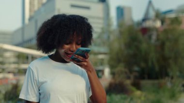 Sapık saçlı bir kadının ağaçlara ve binalara karşı duran sesli mesajını kaydettiği portre. Mutlu Afrikalı Amerikalı kadın, güneşli şehir caddesinde online sohbette iletişim kuruyor.