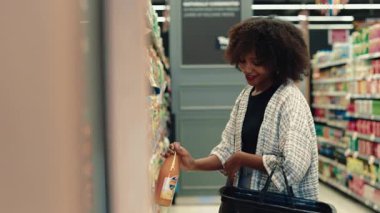 Süpermarket rafından meyve suyu alan gülümseyen Afrikalı Amerikalı kadının portresi. Gevşek ceketli neşeli kadın markette organik içecekleri seçiyor. Sağlıklı beslenme alışkanlıkları