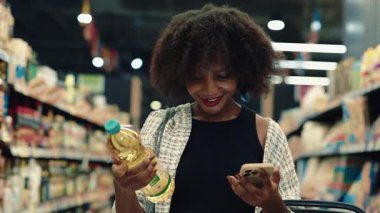 Süpermarketteki bitki yağını kontrol etmek için mobil uygulama kullanan mutlu Afrikalı Amerikalı kadının portresi. Genç bir kadın marketteki organik ürünlerin besin değerini kontrol ediyor. Alışveriş günü.
