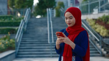 Park merdivenlerinde mesajları okurken gülen Müslüman kadının portresi. Kırmızı tesettürlü güzel Malezyalı kadın şehir caddesinde akıllı telefon kullanıyor. Genç yetişkin yaşam tarzı