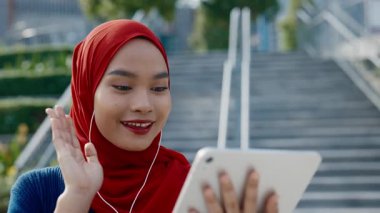 Malezyalı mutlu bir kadının tablet bilgisayarla taş merdivenlere karşı görüntülü aramada partnerleriyle konuşması. Türbanlı kadın, meslektaşlarını güneşli caddedeki çevrimiçi toplantıda karşıladı