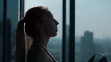 Kadın silueti, salonda panoramik pencereye karşı koşu bandında yürüyor. At kuyruklu kadın, yoğun idmandan sonra kulüpte kardiyo rutininin keyfini çıkarırken vücudundaki gerginliği alıyor. Kapat.
