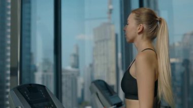Spor salonunda panoramik manzaralı, koşu bandında yürüyen kendine güvenen bir kadının portresi. Uzun at kuyruklu atletik kadın kardiyo egzersizi sırasında şehir manzarasından zevk alır.