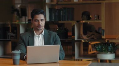 Odaklanmış Hintli erkek girişimcinin portresi şirket ofisinde dizüstü bilgisayarda çalışıyor. Elinde kahve fincanı olan ciddi bir genç adam masadaki e-postaları kontrol ediyor. İş kariyeri