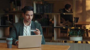 Genç Hintli iş adamının portresi şirket ortamında dizüstü bilgisayarla ödeme yapıyor. Pozitif adam yöneticisi şirket ofisindeki masada otururken online bankacılık hizmetini kullanır.