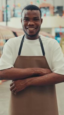 Afro-Amerikan bir adamın portresi. Yemek salonunda kameraya bakarken kollarını kavuşturan geniş bir gülümsemesi var. Üniformalı erkek servis temsilcisi müşterileri açık hava restoranına davet eder. Dikey çekim