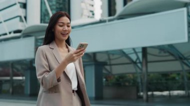 Bej takım elbiseli Asyalı iş kadını şehir ortamında sesli komut özelliği olan bir akıllı telefon kullanıyor. Kullanım sırasında kız mutluluğu ve tatmini.