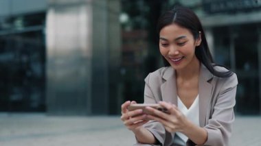 Akıllı telefondan video izlerken muzaffer jestler gösteren Taylandlı iş kadınının portresi. Asyalı kadın şehir ortamında otururken çevrimiçi spor yayını yapmaktan hoşlanıyor