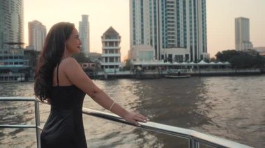 Siyah kokteyl elbisesi içindeki genç Latin Amerikalı kadın portresi yat güvertesinde duruyor. Güzel İspanyol kadın modern şehir manzarasına karşı lüks motorlu teknenin krom korkuluklarına yaslanıyor. Arka plan