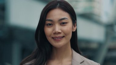 Kameraya bakan gülümseyen Asyalı kadının portresi. Şehir caddesinde dikiliyordu. Neşeli bayan çalışanlar şehir ortamında olumlu tutum sergiliyor. Kurumsal işçi tarzı