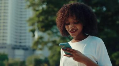 Şehir parkındaki akıllı telefondan internete kayan neşeli Afro-Amerikan kadın portresi. İlgili kadın, şehir ortamında cep telefonuyla sosyal medya içeriğinden hoşlanıyor