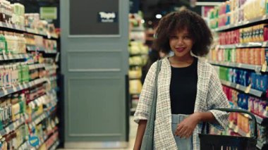Süpermarket koridorunda elinde alışveriş sepeti tutan Afro-Amerikan kadının portresi. Günlük giysiler içinde neşeli bir kadın marketten yiyecek seçiyor. Boşluğu kopyala