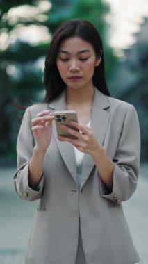 Taylandlı iş kadınının portresi şehir merkezinde telefonu tutarken yumruğunu zaferle gösteriyor. Takım elbiseli Asyalı kadın başarılı proje tamamlanmasıyla ilgili harika haberler alıyor. Dikey video