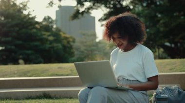 Green Park 'ta modern bir laptopta gülümseyen bir kadının portresi. Genç Afrikalı Amerikalı bayan internette çimlerin üzerinde oturuyor. Uzak görev