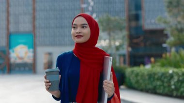 Kahve fincanı ve dizüstü bilgisayarla şirket binasının önünde yürüyen Müslüman kadın ofis çalışanı portresi. Şehir merkezindeki profesyonel şirket çalışanı. Kariyer fırsatları dahil