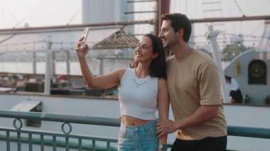 Genç Latin Amerikalı kadın ve erkeğin portresi turist gemisinin yanındaki akıllı telefondan selfie çekiyor. Mutlu İspanyol turistler tatil köyü limanında poz veriyor. Birlikte romantik bir tatil.