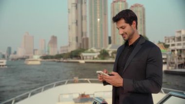 Genç İspanyol iş adamının portresi yatta duran akıllı telefonu tutuyor. Pozitif Latin girişimci döviz kurlarını kontrol ediyor. Turist motorbotu gökdelenlere karşı.