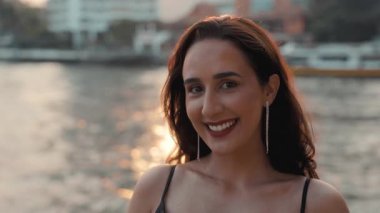 Neşe dolu bir Latin Amerikalı kadının portresi geniş bir kesime gülümseyerek kıyıdaki sakin koy suyuna karşı duruyor. Esmer İspanyol kadın gün batımında şehir setinde kameraya bakıyor.