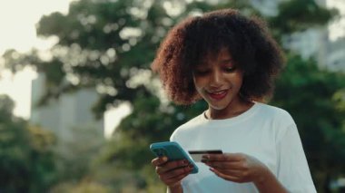 Yaz parkında kredi kartıyla ödeme yapan güzel Afrikalı Amerikalı bir kadının portresi. Kadın kamu bahçesinde akıllı telefondan mobil uygulama ile ödeme yapıyor