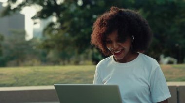 Şehir parkında laptopa el sallayan dost canlısı bir kadının portresi. Neşeli Afrikalı Amerikalı bayan yaz bahçesinde arkadaşlarıyla ve iş arkadaşlarıyla görüntülü sohbet ediyor.