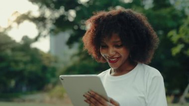 Yeşil yaz parkında elinde tablet bilgisayar tutan Afro-Amerikalı bir kadının portresi. Genç bir kadın, şehir caddesinde dijital cihazla görsel içeriği izliyor. Şehir tarzı