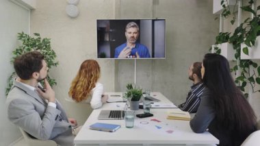 Bir grup ofis çalışanı bir video konferans toplantısına katılıyor, ekranda bir erkek konuşmacıyı izliyorlar. Tablolar ve dizüstü bilgisayarlarla dolu bir iş ortamı. Erkek akıl hocası konuşuyor.