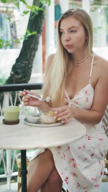 Yuvarlak bir mermer masada oturmuş Tayland usulü hindistan cevizi sütlü pilav yiyen ve tropik bir kafede matcha latte yudumlayan genç bir kadının dikey portresi.