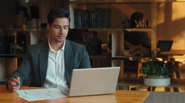 Hintli mimar portresi, iş yerinde tasarım ve dizüstü bilgisayarla çalışıyor. Odaklanmış mühendis çalışma odasındaki ahşap masadaki ev çizimlerini kontrol ediyor. İnşaat bürosu.