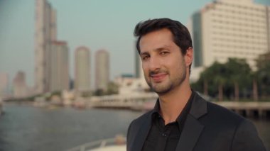 Takım elbiseli Latin Amerikalı işadamının portresi yat güvertesindeki kameraya gülümsüyor. Mutlu İspanyol adam şehir merkezindeki gökdelenlere karşı modern motorlu teknede dinlenmeyi seviyor.