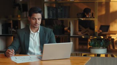 Genç Hintli tasarımcının portresi şirket işyerindeki dizüstü bilgisayarın planlarını kontrol ediyor. Mühendis, birlikte çalışırken bilgisayarla birlikte çalışma masasında bina çizimleri geliştiriyor. Yaratıcı iş