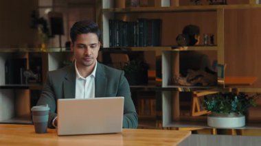 Genç Hintli girişimcinin portresi iş yerinde dizüstü bilgisayarla çalışırken kahve içer. Kendine güvenen serbest çalışan bir adam internet işini masa başında bilgisayarla yürütüyor. Ticari faaliyet