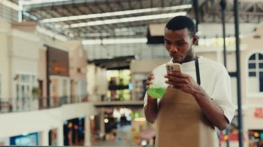 Alışveriş merkezinde içki içerken elinde telefon tutan Afro-Amerikan bir adamın portresi. Erkek garson teneffüste sosyal medyayı kontrol ediyor. Servis elemanı ferahlatıcı içecekten hoşlanıyor.